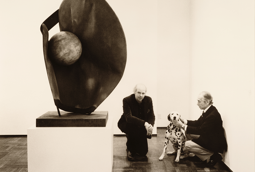 Two older white men sit with a dalmatian next to a sculpted art piece on display