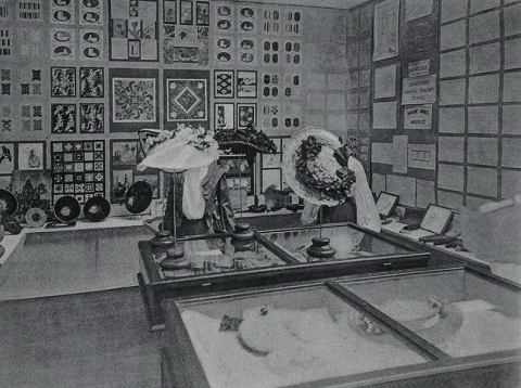 Photo shows items made by students. Walls are filled with samples of paintings, needlework, and reports. Bowls are displayed on a counter. Hats on stands sit on glass-topped cabinets.