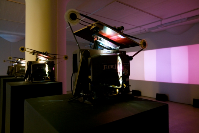 Four film projectors in a darkened gallery project color fields in a superimposed fashion.