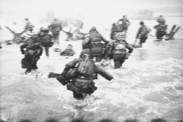 Click to view larger image A grainy black and white image of troops walking through ocean water.