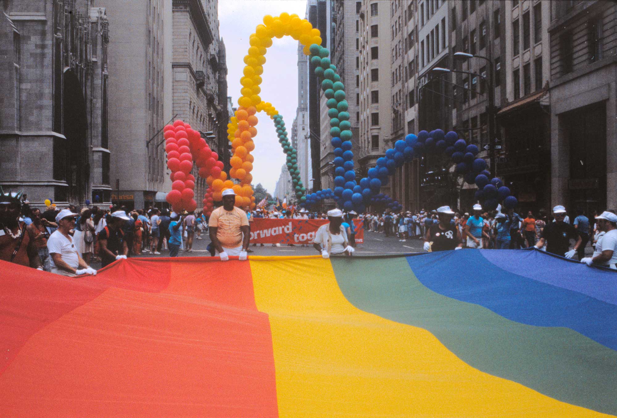 Group of people holding a rainbow-striped banner across a city street. There is a parade behind them.
