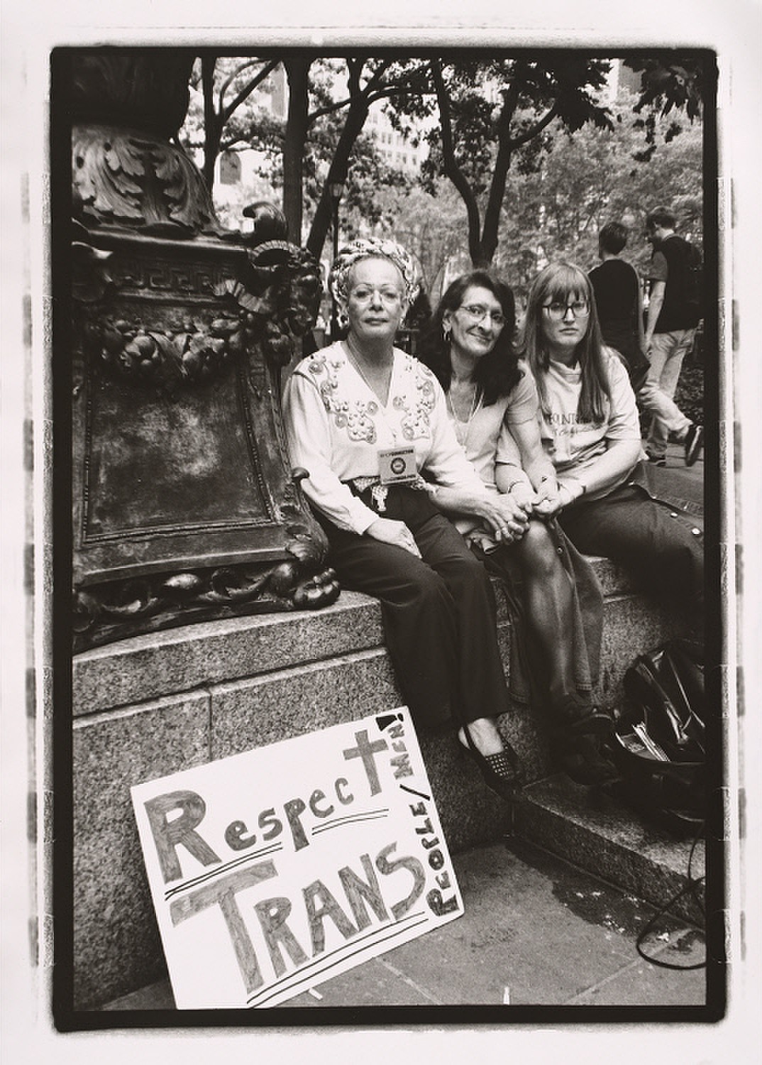 Sylvia Rivera sitting between two women on an outdoor stone wall. A sign at their feet reads "Respect trans people/men!"