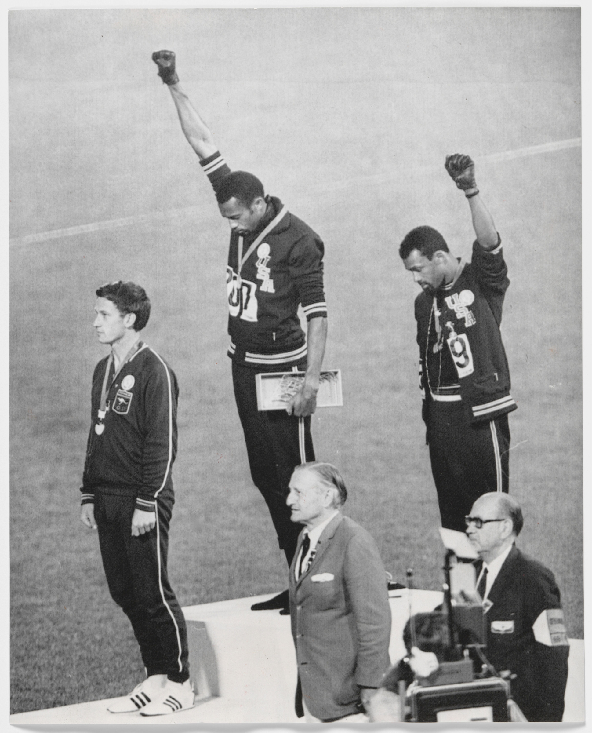 Tommie Smith, John Carlos and Peter Norman stand on an Olympic podium. Smith and Carlos have their fists raised.