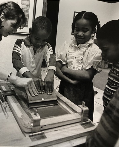 In a black-and-white photo, an adult helps a child pull a squeegee across a silkscreen frame to make a print while other children watch