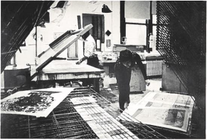 In an art studio, Lou Stovall lifts a silkscreen frame while Di Stovall places a print on a rack to dry. Visible on a different rack in the foreground is a silkscreened poster for a Roberta Flack concert.