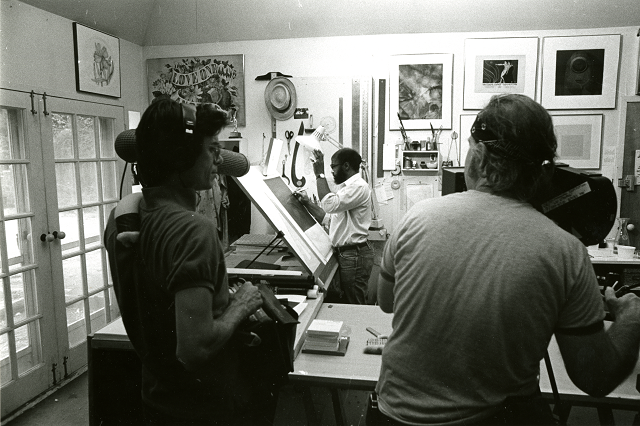 An artist stands while making stencils for silkscreen printing. In the foreground, two people record the artist for a film. One holds a microphone and sound recording equipment while another operates a 16mm camera on a tripod. Artwork hangs on the walls, and light enters through the glass panes of French doors in front of the artist's upright drafting table.. 
