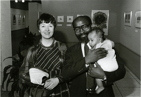 A white cloth is draped over a woman's arm as she stands next to her spouse, who holds their baby. Framed artworks are visible on the gallery walls behind them in this black-and-white photograph.