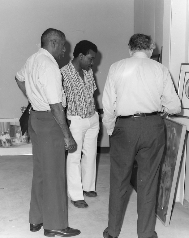 John J. Robinson, David Driskell, and Joshua Taylor look at framed works of art leaning against a wall in this black-and-white photo.