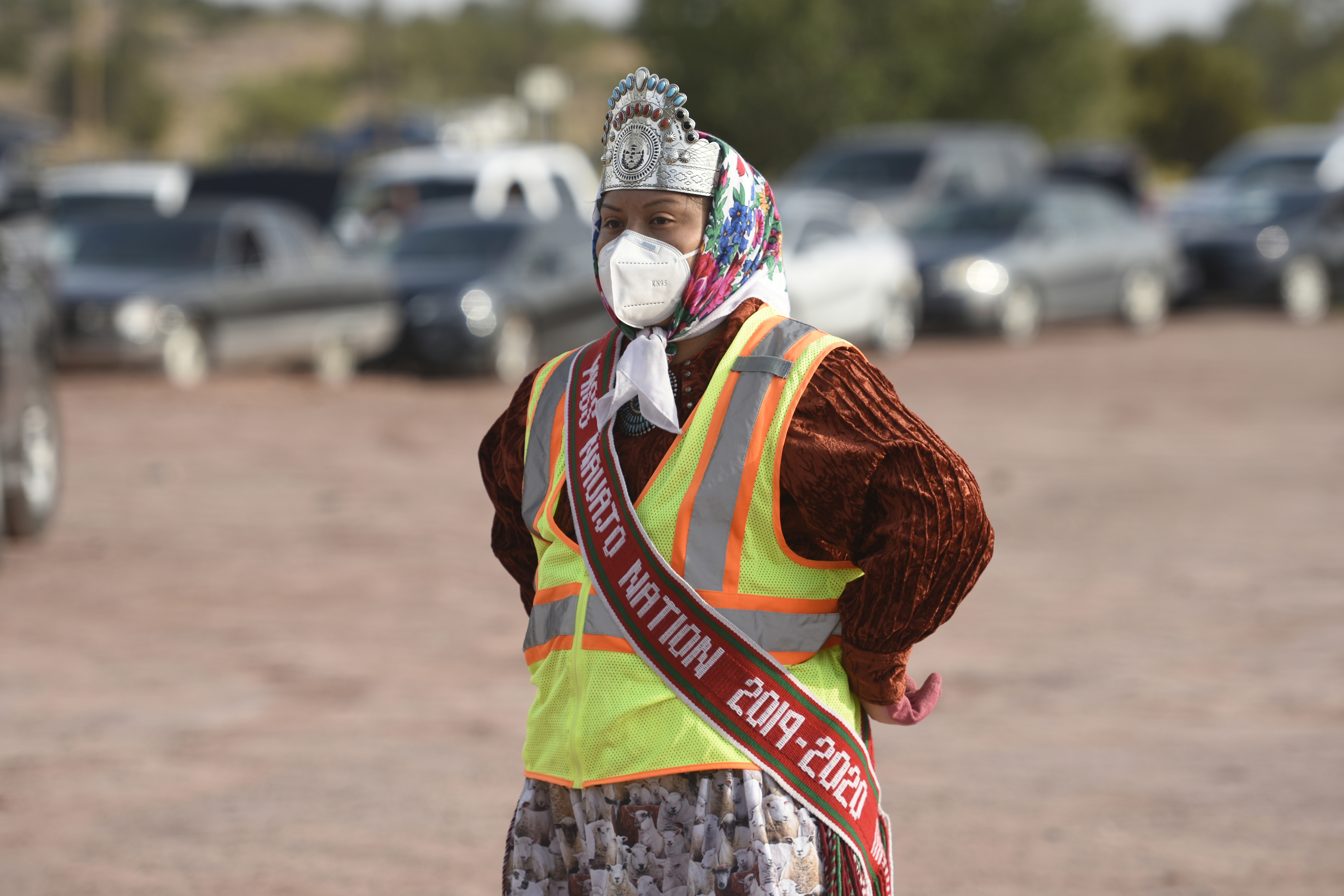 Woman wearing safety gear, headdress, and sash stands in a dirt field with cars in background.