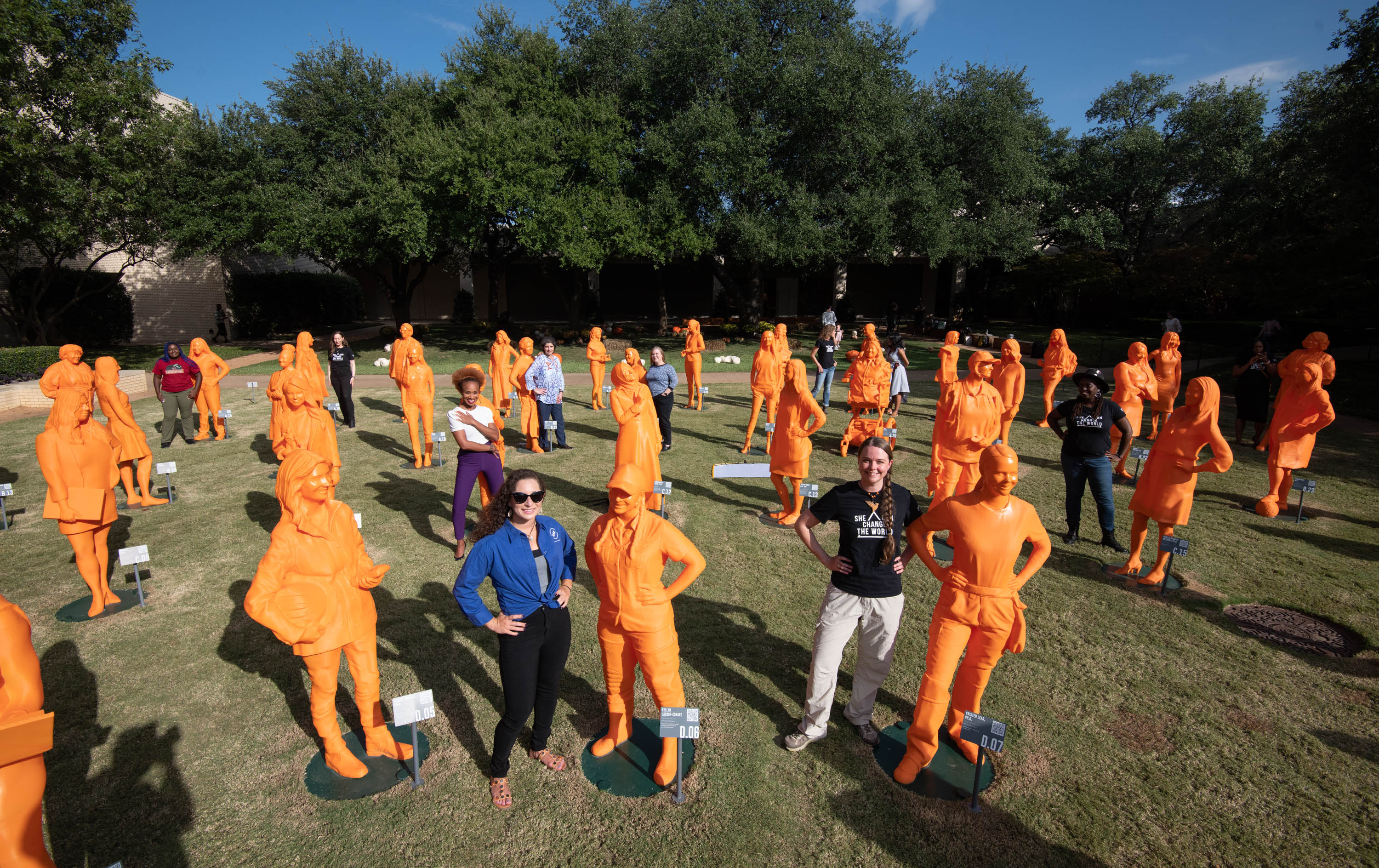 Group of orange, women statues in a open field along with two real humans