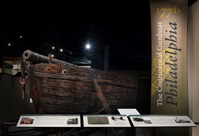 Museum gallery with three text panels in front of a large wooden boat that is clearly aged.