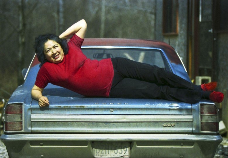 Woman in red shirt and shoes poses on hood of car