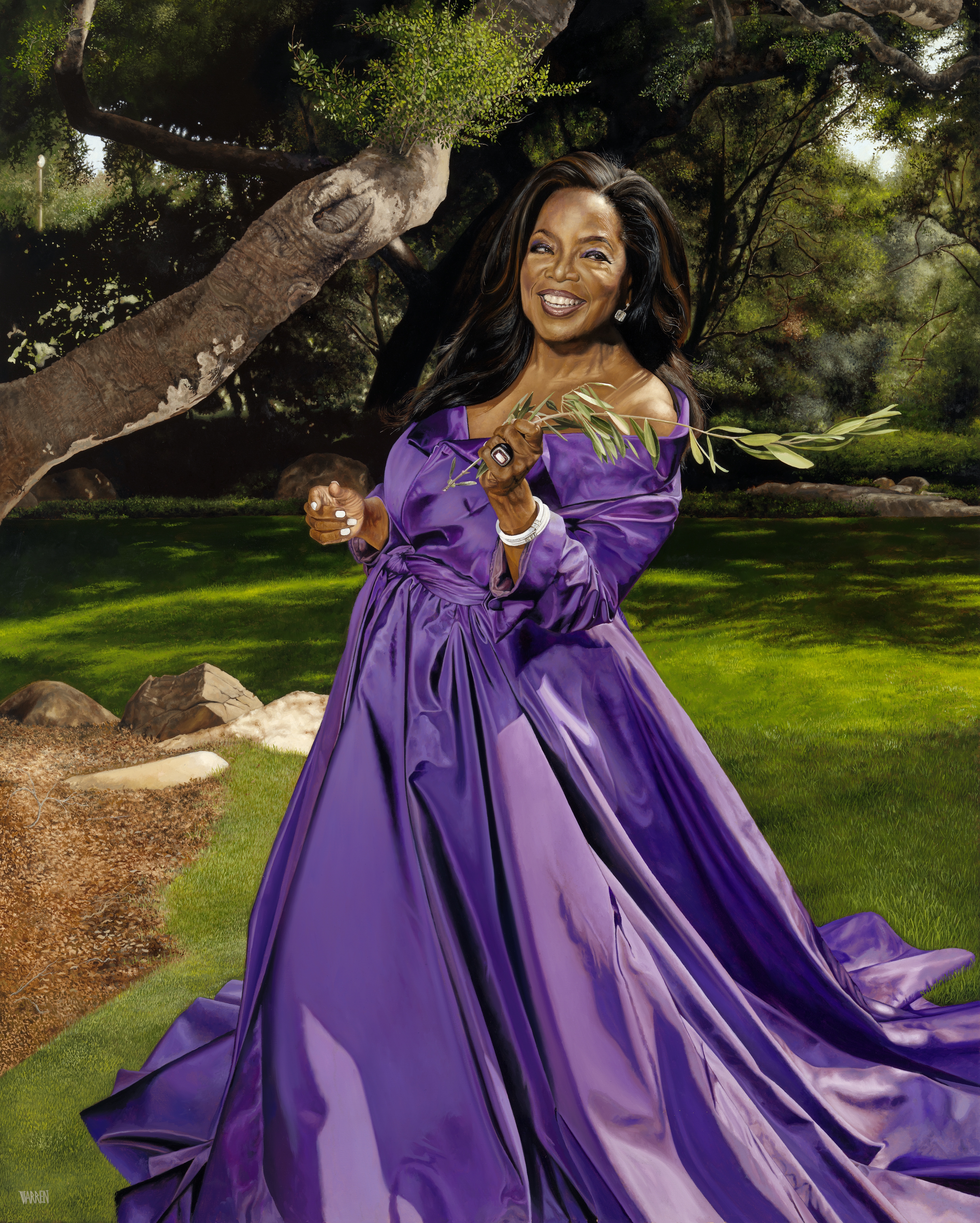 Woman in long, silky purple dress holds an olive branch while standing in a garden with a large tree branch in the background.