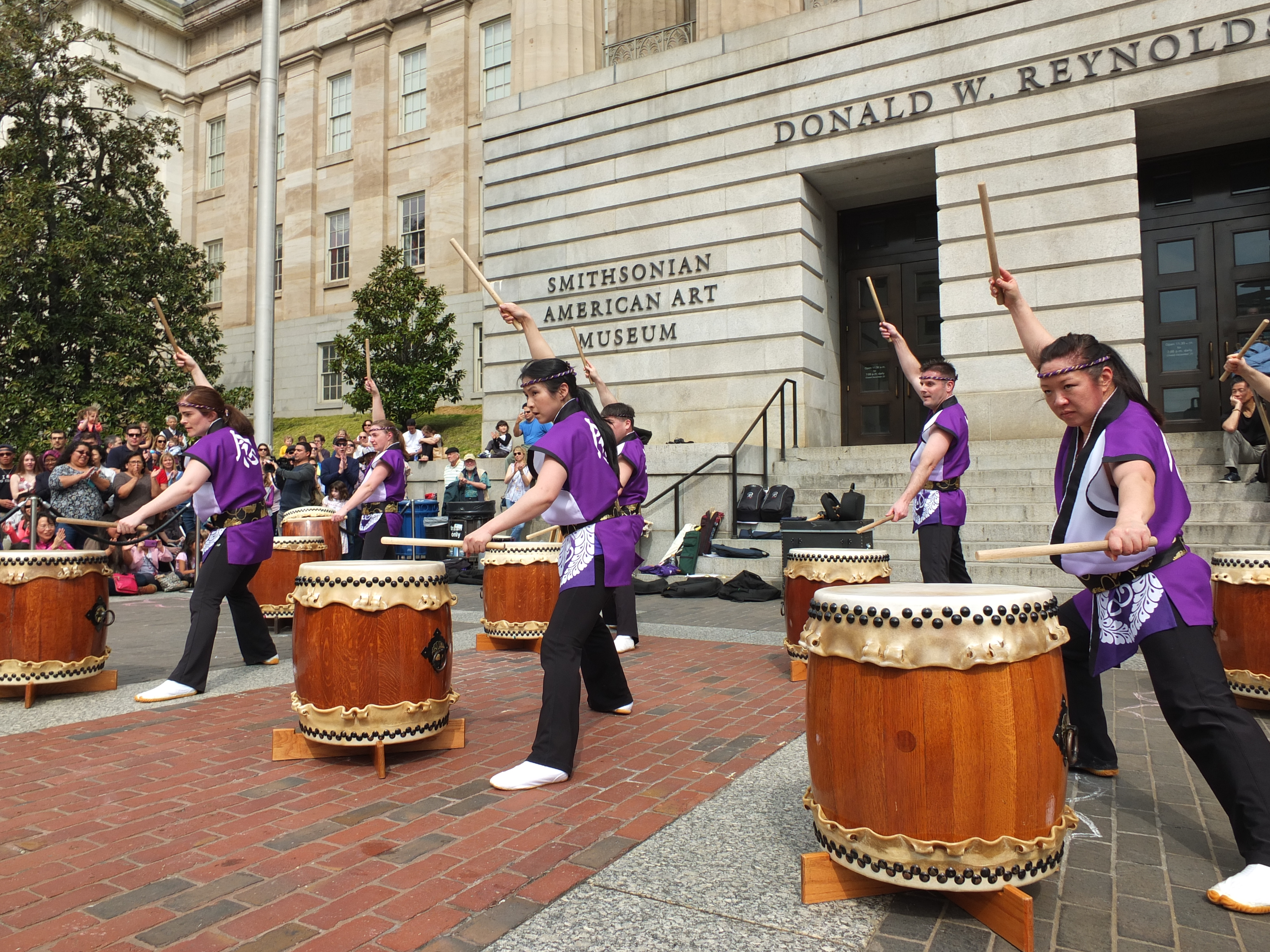 Group of large drums are being played by drummers outside American Art Museum