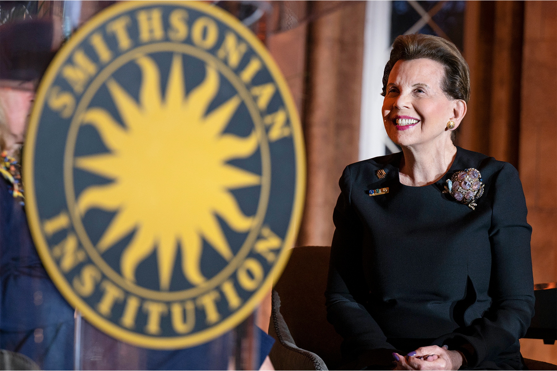 Adrienne Arsht wearing a black, long-sleeved dress with bejeweled frog broach standing next to the Smithsonian logo