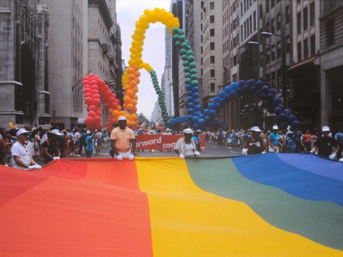 Group of people holding a rainbow-striped banner across a city street. There is a parade behind them.