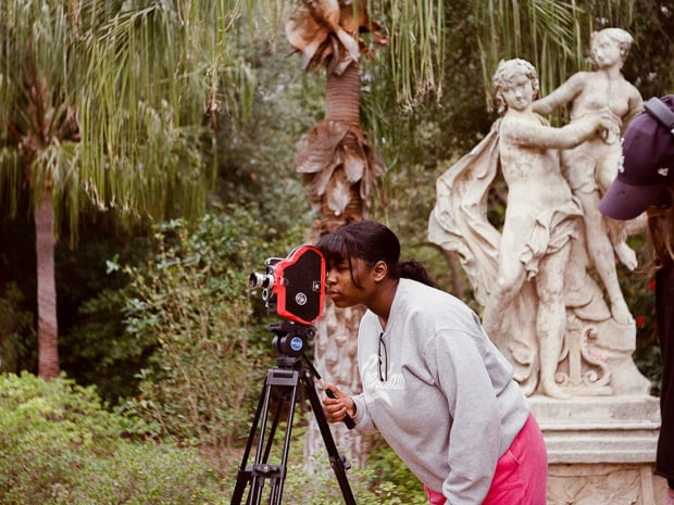 A girl in a light grey sweatshirt looks into large camera on tripod in outdoor sculpture garden. 