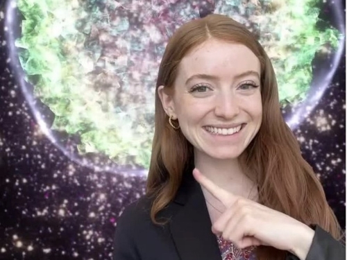 Girl with red hair points at graphic projection surrounding her, which looks like black starry sky with large neon green orb.