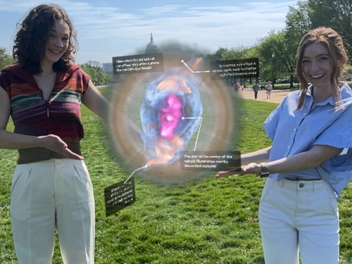 Two women stand on a grassy area pointing with their hands toward graphic projection of a starry nebula between them.