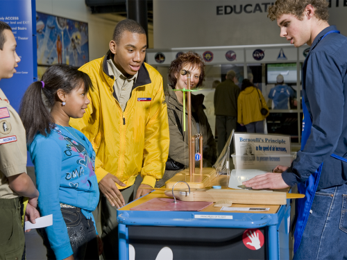 A young girl in a blue top stands next to a taller man in a yellow jacket in front of a rolling cart demonstration.