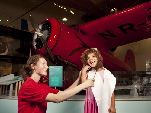 Museum Explainer Rose Agger helps a young girl dress up like a historic aviator