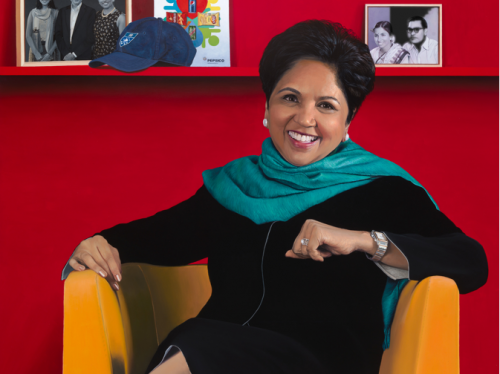 Indra Nooyi by Jon R. Friedman sitting in an orange chair with a red wall behind her.