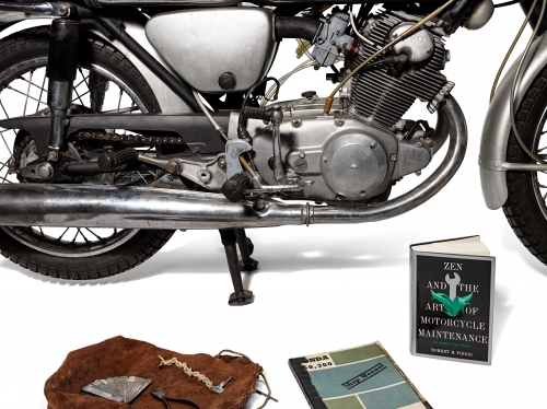 Black motorcycle with two books and a leather bag on ground next to it.