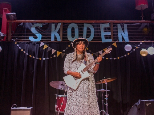 Female guitarist on stage, "Skoden" banner in background