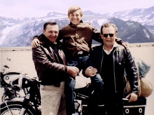 Two men in jackets hold up a young boy between them and smile at the camera, standing in front of motorcycle.