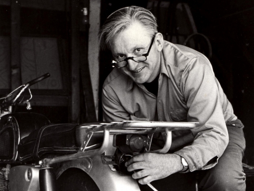 Man wearing glasses and long-sleeved button-down with pants leans over back tire of motorcycle.