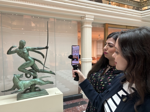 Two woman with long, dark hair stand in front of small sculpture, holding phone up toward the sculpture.