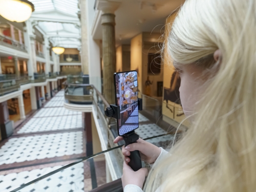 Girl with blond hair stands on interior balcony overlooking large space and holds her phone with an AR graphic on it.