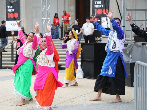 Picture of people outside in front of marble steps, wearing layers of colorful clothes dancing with their arms up