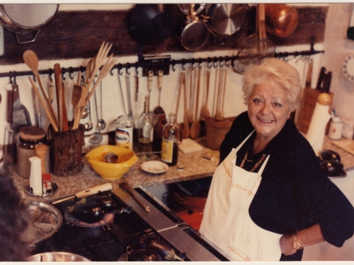 A woman with short white hair, black top and white apron stands on a kitchen with a stove and various utensils.