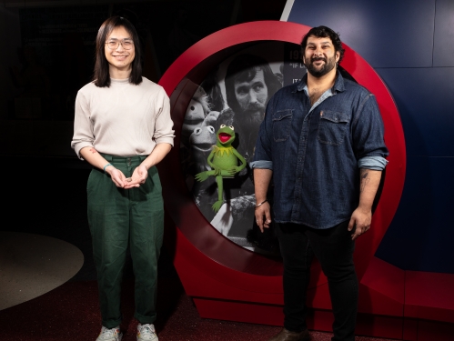Two men stand beside green felt puppet, Kermit.
