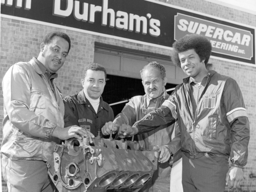 Black and white photo of four men grouped around an auto engine
