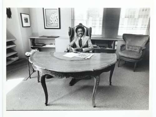 Black and white photo of Byron Lewis, sitting at half-moon-shaped desk