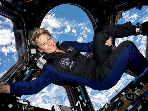 Woman in navy jumpsuit floats in spacecraft with windows showing upper Earth atmosphere.