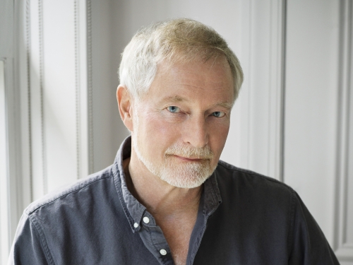 Man with short white hair, wearing a dark gray button down, sits casually with arm propped on leg