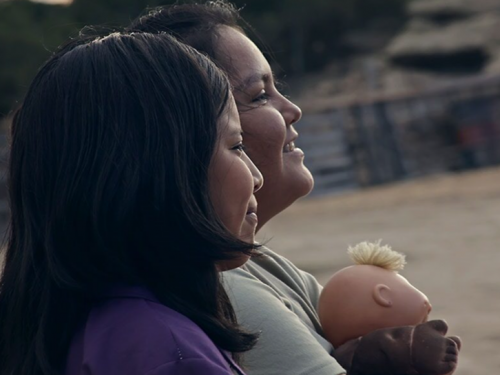 Profiles of two women with dark hair, smiling, as one carries a doll toy.