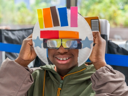 Young boy with braces smiles as he wears reflective sunglasses and paper mache hat.