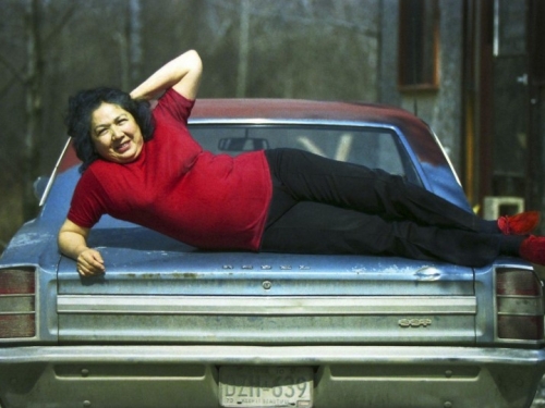 Woman in red shirt and shoes poses on hood of car