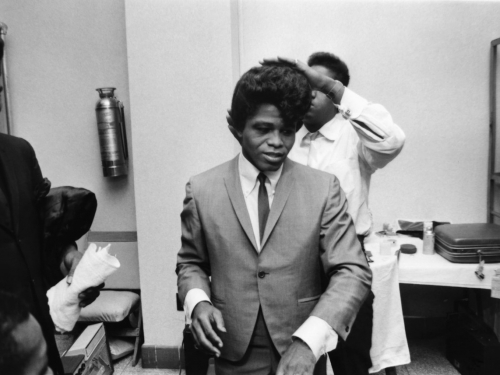 Black and white backstage image of singer James Brown in a suit, with a person behind him running their hand over James's hair