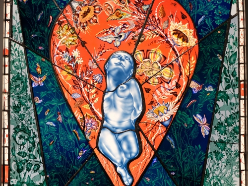 Image of stained glass in various colors with stylized figure floating in center with hands behind its back, looking upward.