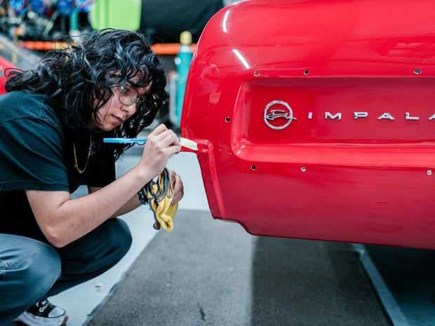 A young person wearing jeans and short sleeve shirt crouches next to end of red Impala. 