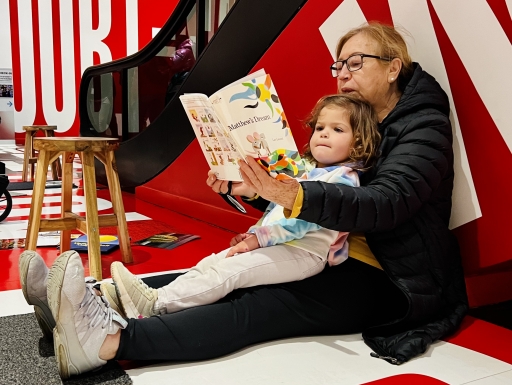 A grandparent reads her young granddaughter a story on the floor of an art museum.