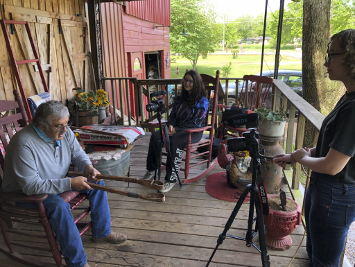 A community leader tells stories on a porch while a filming crew captures the moment.