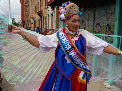 Color photo of a woman in a parade with extended arms holding pleated, iridescent wings with halter neck.