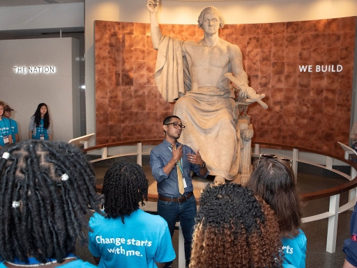 An educator stands in front of a George Washington statue addressing a group of students in bright blue matching t-shirts