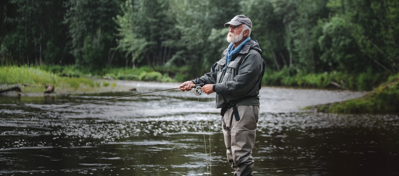 Man with white beard wearing waders fishing in stream 